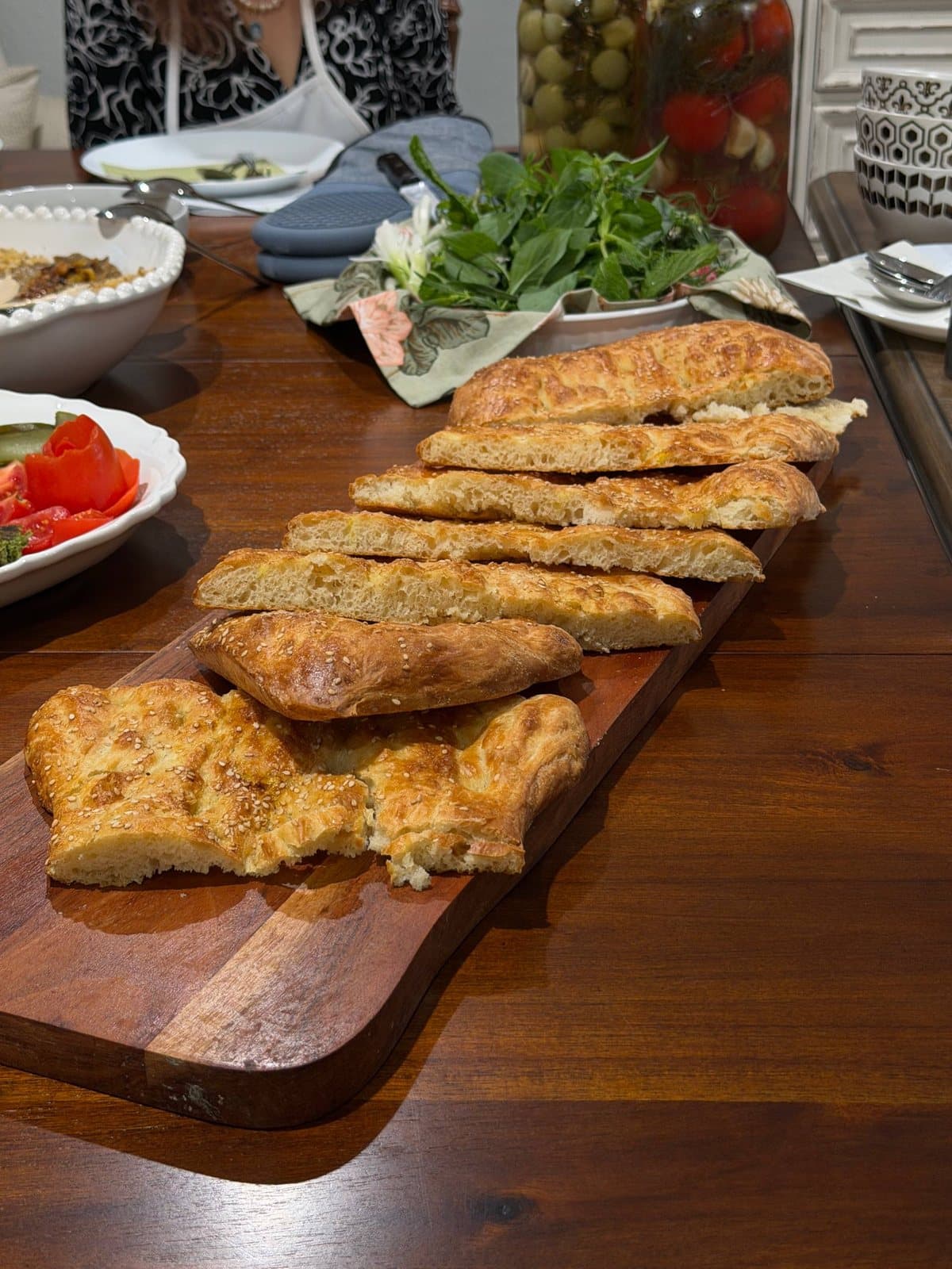 Sesame barbari sliced along a long board, beside fresh herbs, pickles and tomatoes